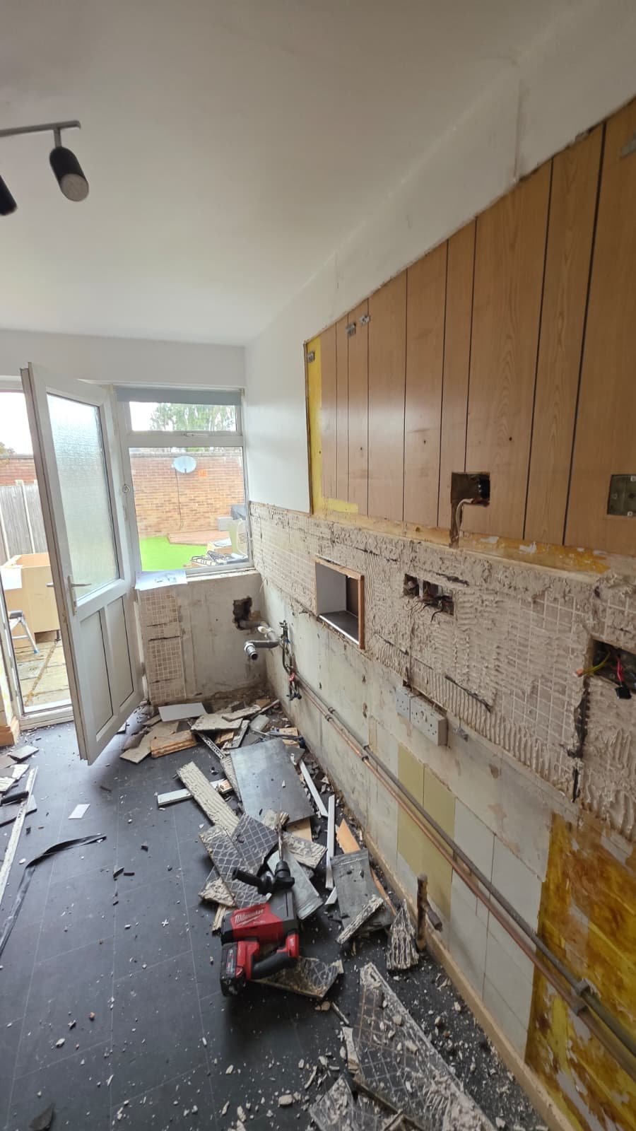 Kitchen undergoing renovation with stripped walls, debris on floor, exposed wooden cabinets, and door to backyard