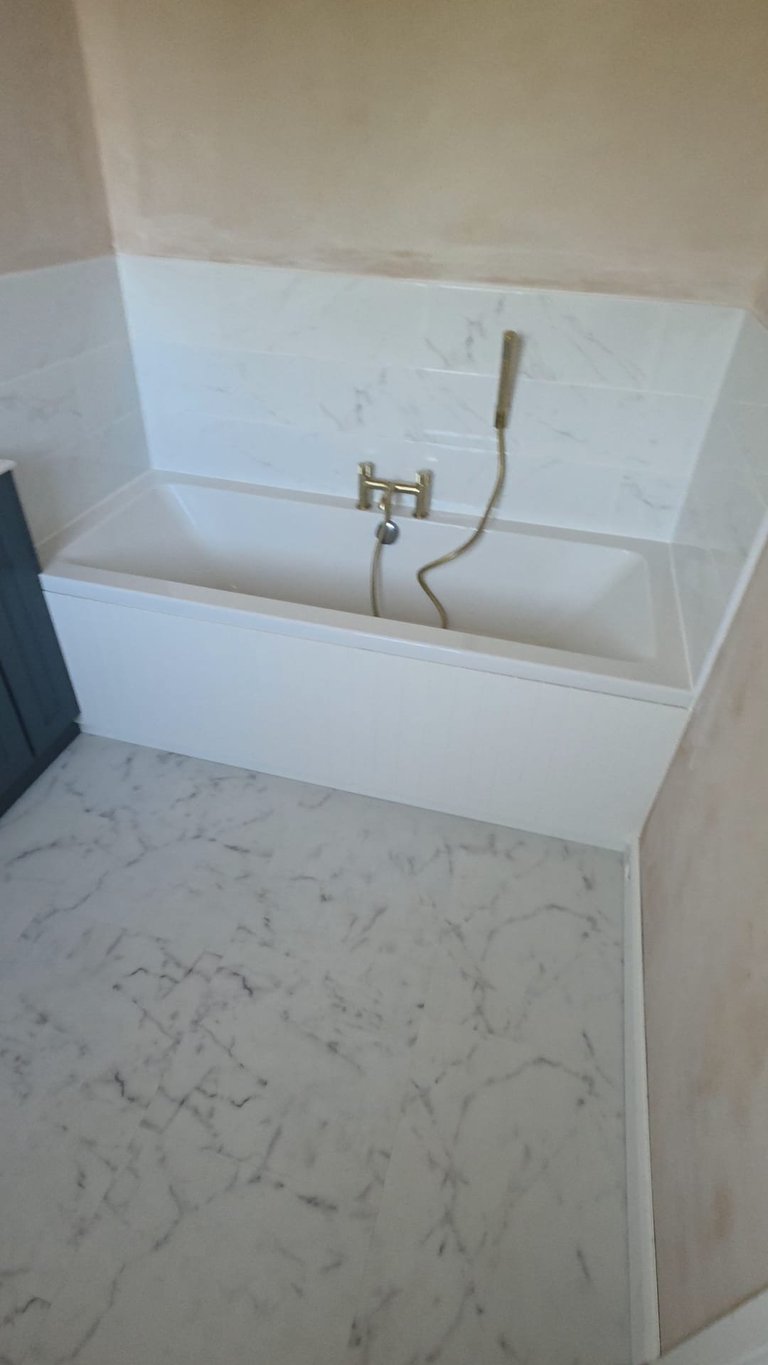 Modern white rectangular bathtub with marble floor and cream walls, featuring brass faucet and handheld shower head attachment
