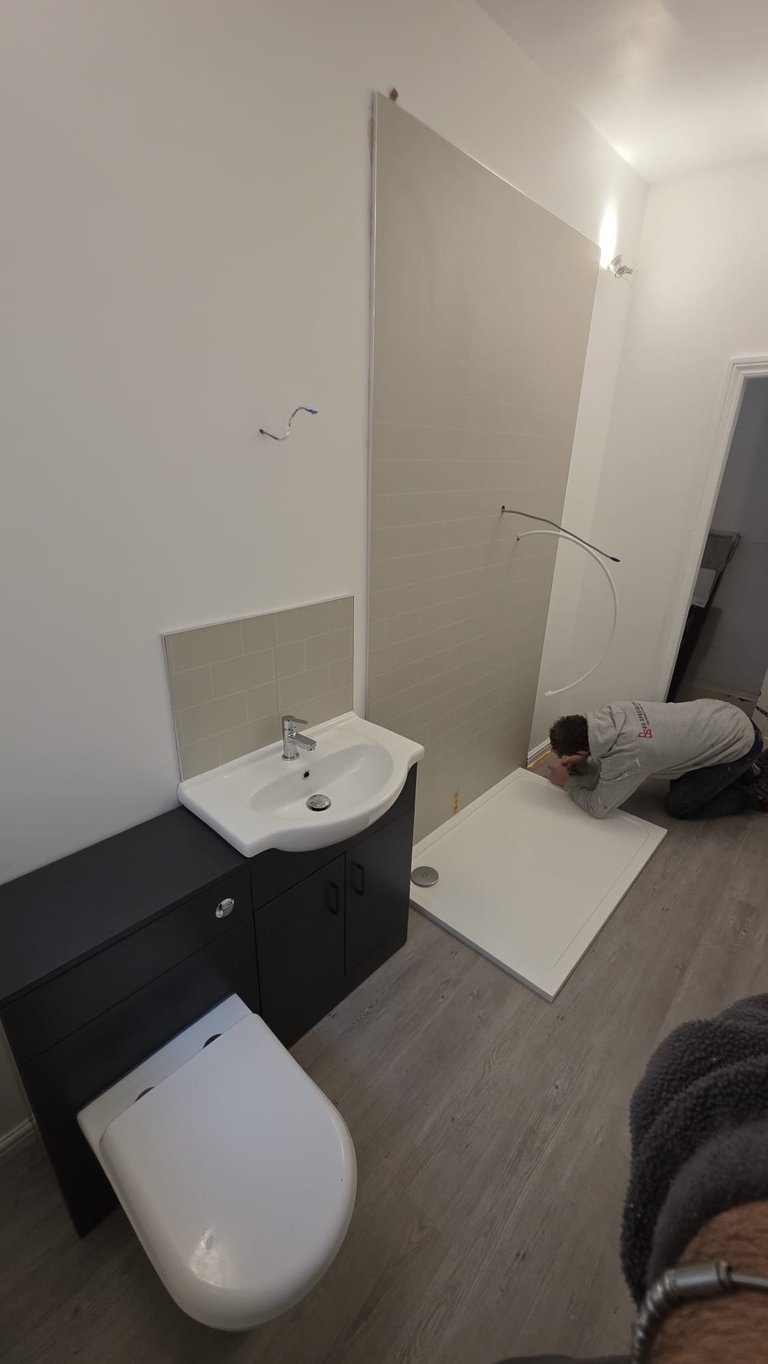Modern bathroom with white sink, black vanity cabinet, white toilet, and a person installing shower enclosure panels