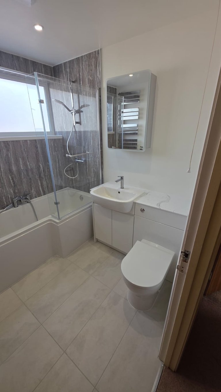 Modern bathroom with tiled flooring, glass shower enclosure with wood-grain tile, white toilet, pedestal sink, and mirrored medicine cabinet