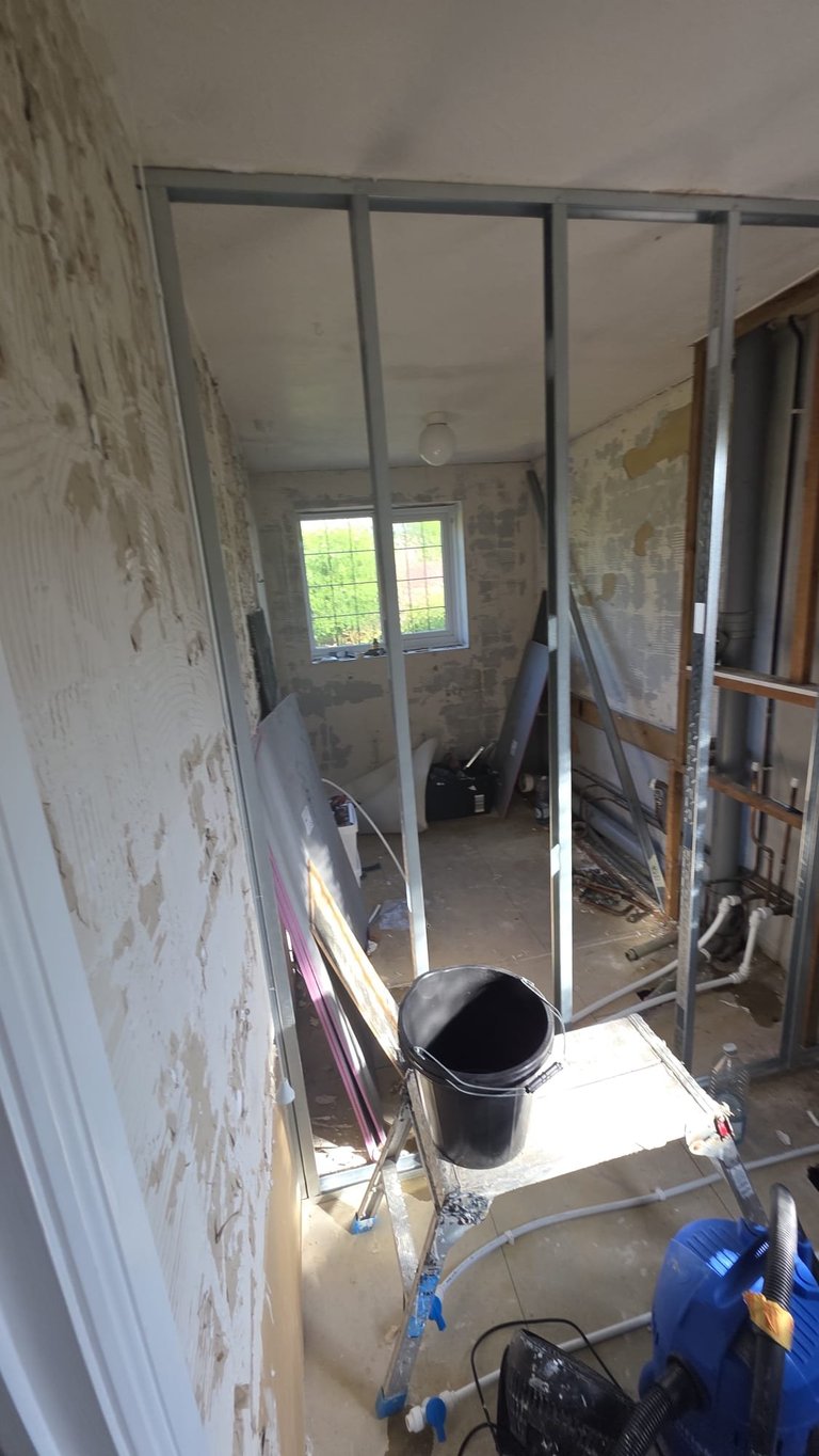 Construction site bathroom with metal framing, glass shower enclosure under renovation, peeling walls, and tools scattered on floor