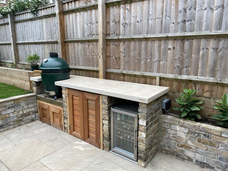 Outdoor kitchen setup with built-in grill, stone countertop, wooden storage doors, and green egg smoker on brick patio