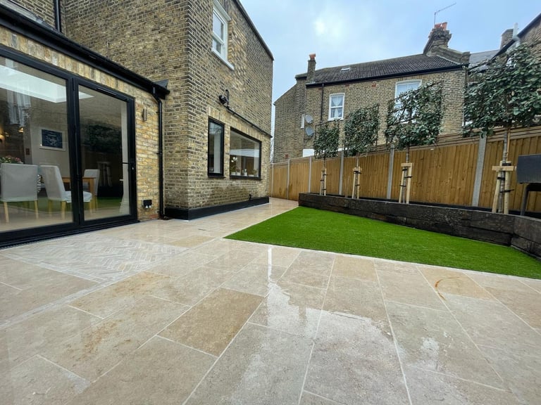 Modern patio with stone tiles, artificial green lawn, wooden fence, and ivy-covered brick houses in background