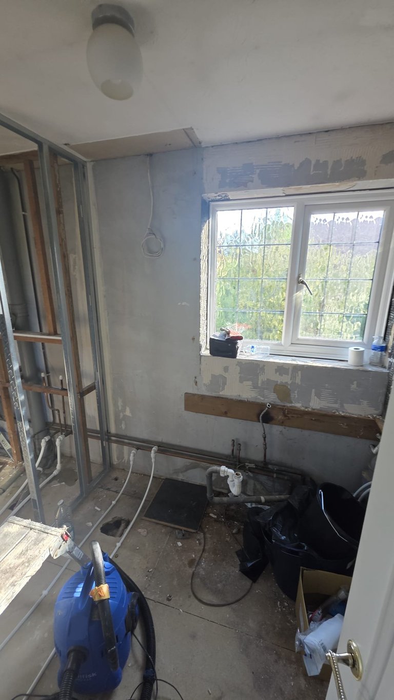 Unfinished bathroom under renovation with exposed drywall, window, glass shower enclosure, and blue cleaning equipment on floor