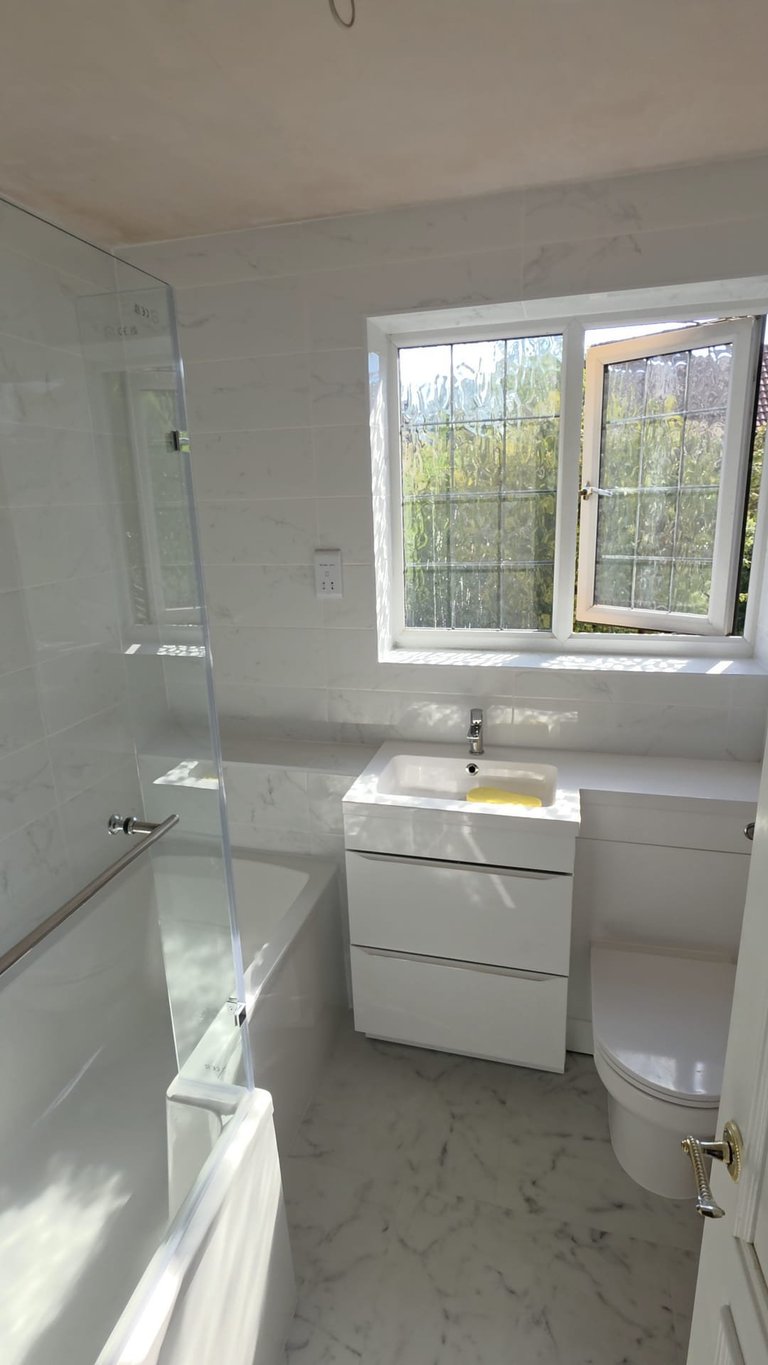 Modern bathroom with white vanity, toilet, glass shower enclosure, and window overlooking green outdoor area
