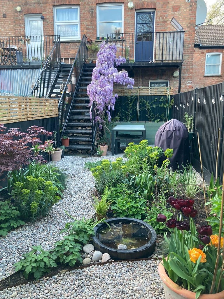 Urban brick courtyard garden with blooming purple wisteria vine, circular water feature, flowering plants, gravel pathways, black metal railings, and external stairs to upper level