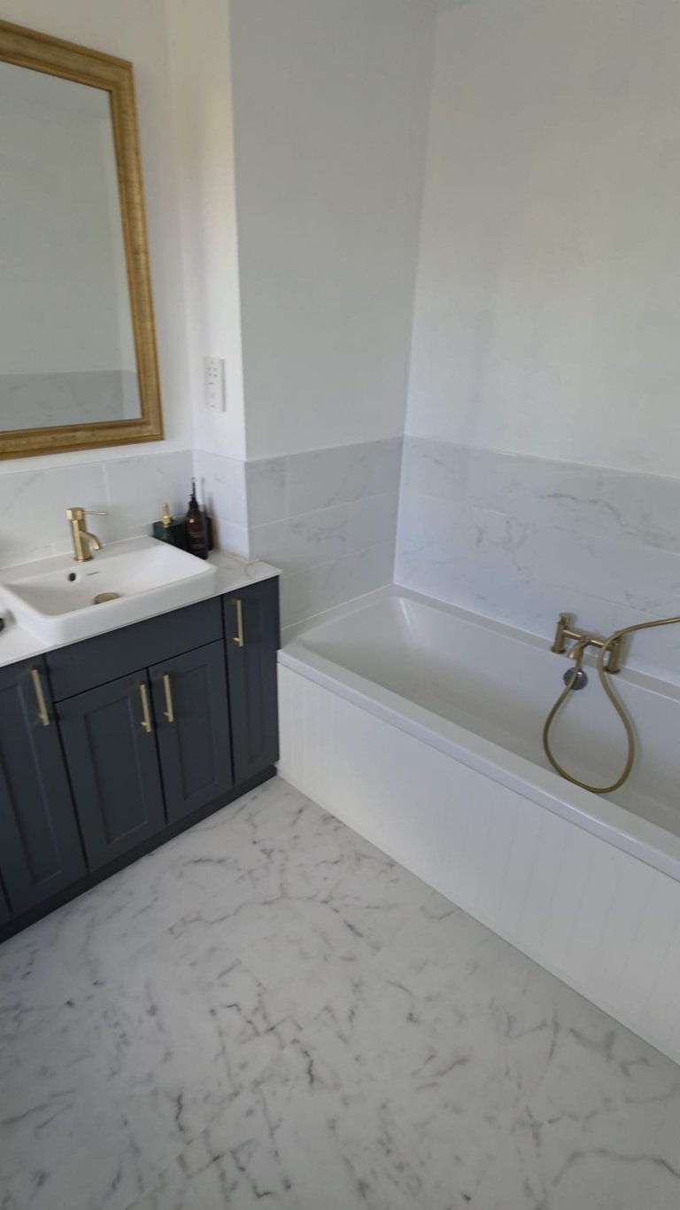 Modern bathroom with marble floor, dark vanity cabinet, white bathtub with brass fixtures, and gold-framed mirror