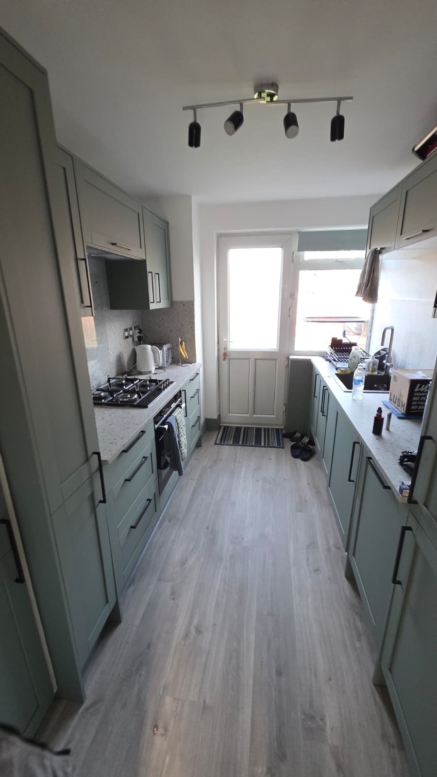 Modern galley kitchen with sage green cabinetry, gray countertops, wood-effect flooring, and black pendant lights overhead